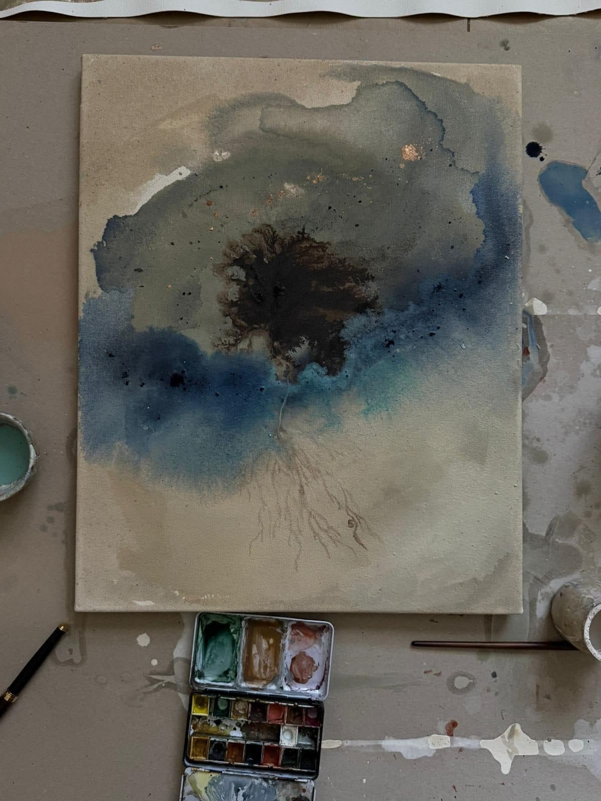 Obra abstracta de Carla Kasseroler en azules y tierras, en el suelo del taller junto a una paleta de acuarelas.