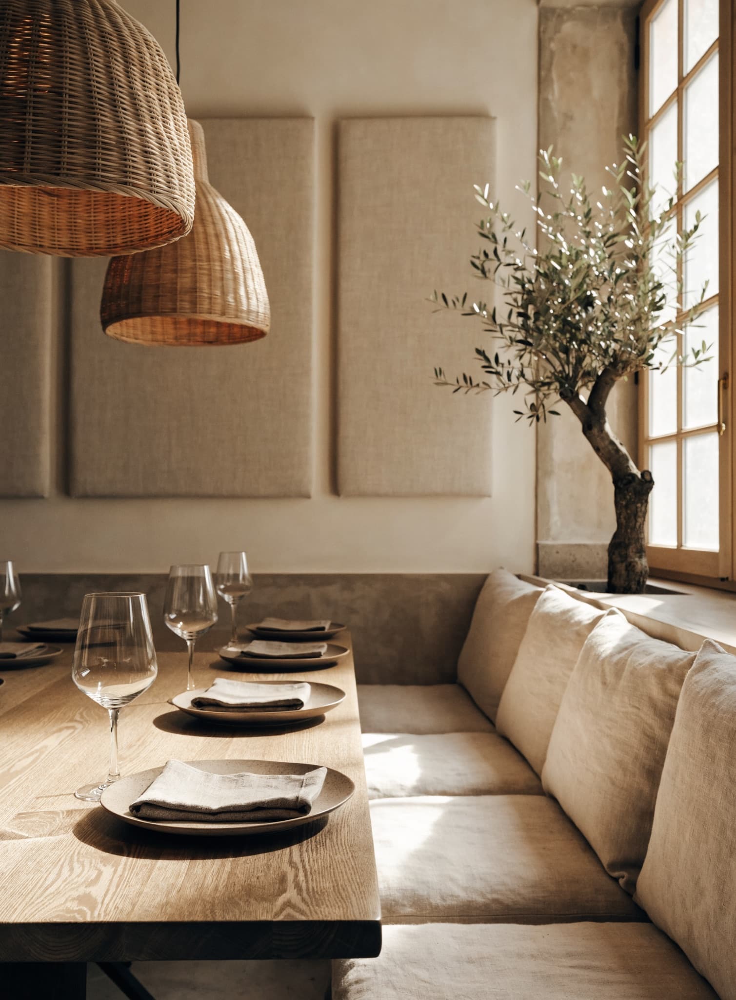 Rincón de restaurante con banco corrido de lino, mesa de roble con copas, dos lámparas de mimbre y tres paneles beige lisos en la pared, junto a un olivo y una ventana soleada.