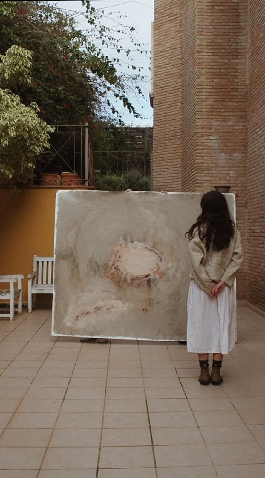 Una mujer de espaldas mira una obra abstracta de Carla Kasseroler apoyada en la pared de un patio.