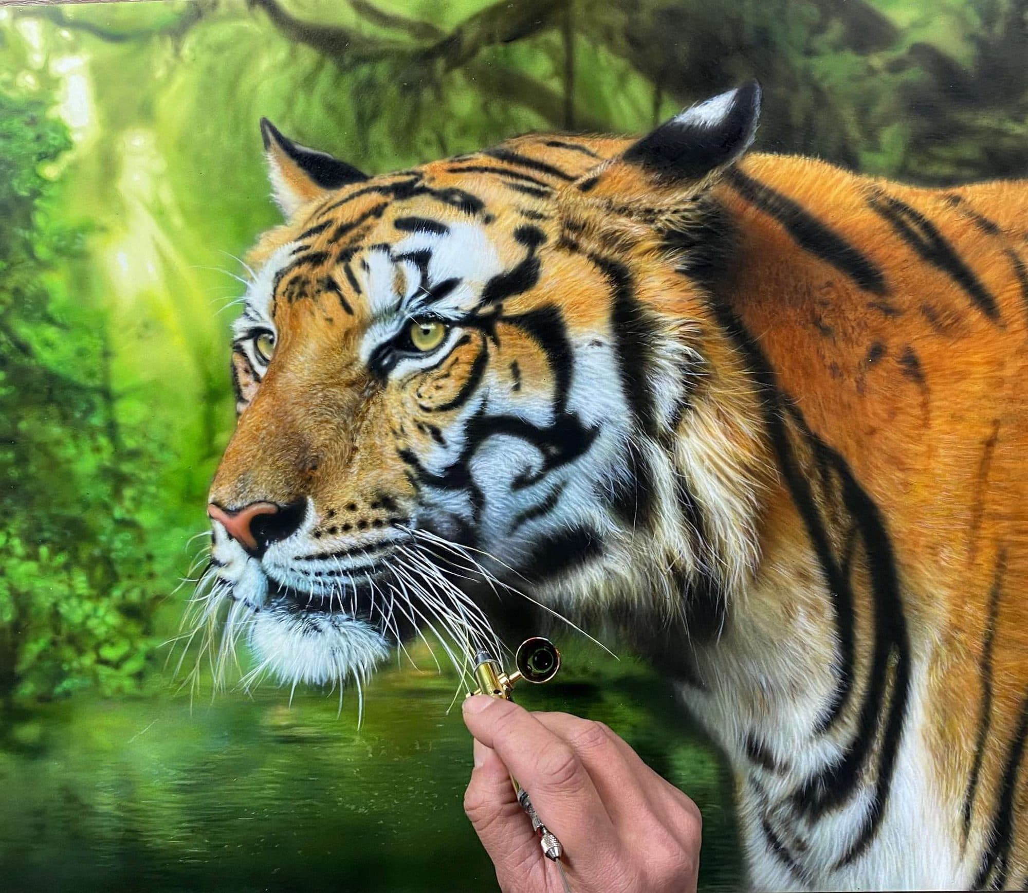 Obra hiperrealista de Marissa Oosterlee: un tigre entre la vegetación, con la mano de la artista y el pincel en primer plano.