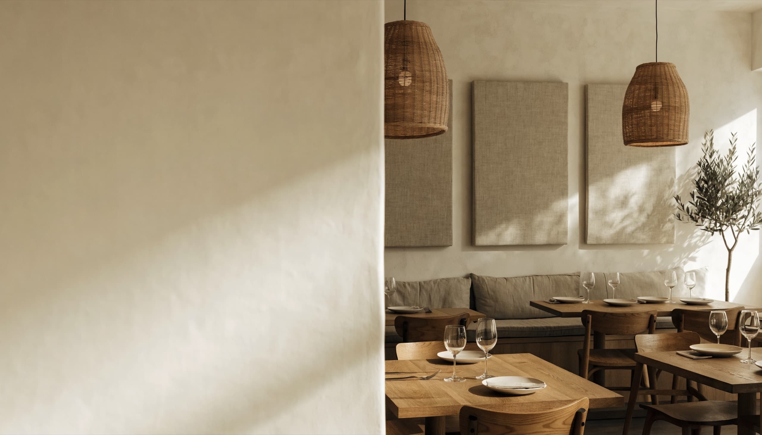 Sala de restaurante con mesas de roble puestas, banco corrido de lino, dos lámparas de mimbre y tres paneles beige lisos en la pared, junto a un olivo; a la izquierda, una pared de yeso vacía con luz cálida.